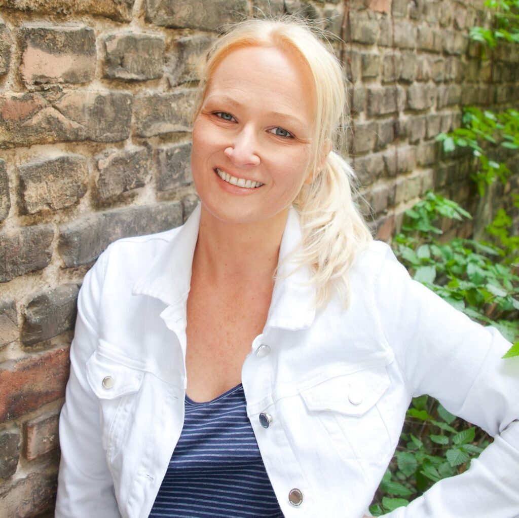 Blonde Frau mit hellem Teint lacht freundlich vor einer alten Backsteinmauer